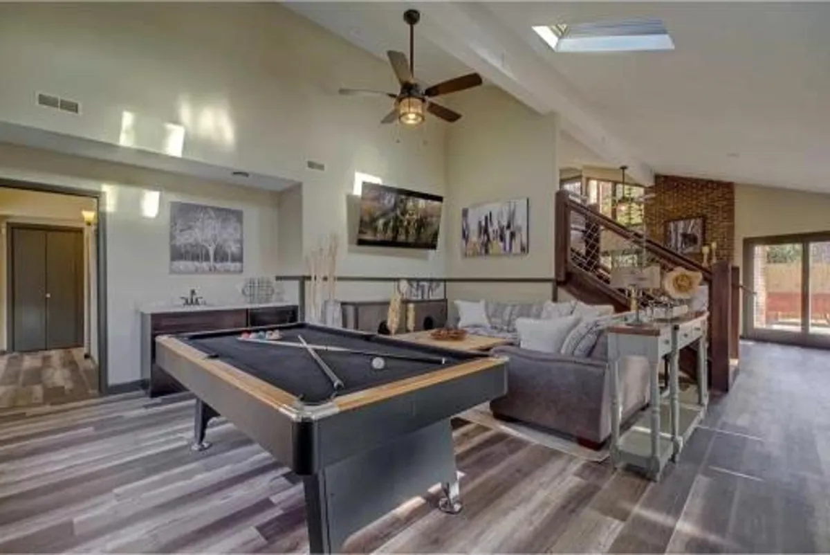 A lively game room featuring a pool table as the central attraction, complemented by a comfortable sofa, vibrant artwork on the walls, and an open staircase. The space exudes a casual, social atmosphere ideal for friendly competition and relaxation.
