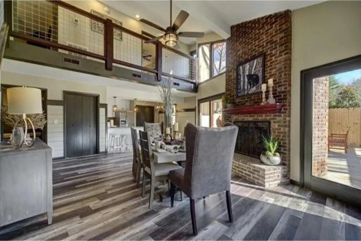 This image showcases a bright, newly remodeled dining area featuring a dining table set for a meal, a classic brick fireplace, and large windows that let in natural light. The modern ceiling fan and subtle décor add a touch of sophistication and comfort to the space. The fireplace and windows create a cozy yet upscale ambiance, perfect for gatherings.