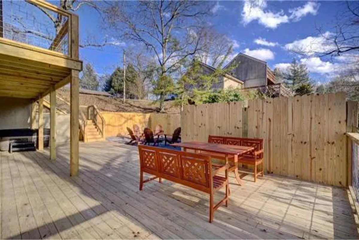 A rustic wooden deck with a cozy seating area overlooking a fenced backyard bordered by lush greenery. The perimeter fence ensures privacy and safety, making it ideal for both adult relaxation and children's play.