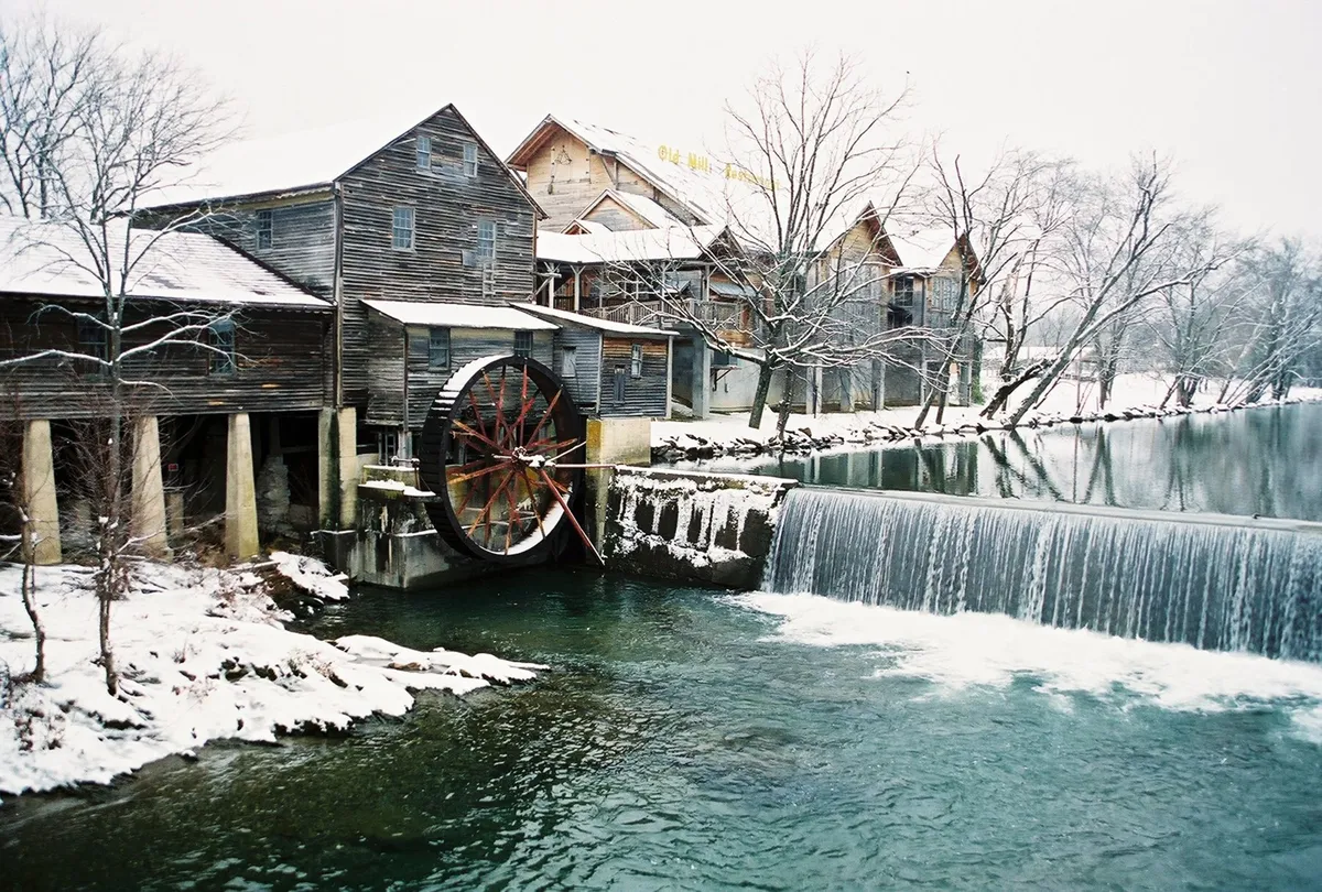A historic waterwheel sits beside gently flowing water, with snow-covered buildings and trees creating a peaceful winter scene. The landscape offers opportunities for quiet walks, historical exploration, and enjoying the tranquil side of nature.