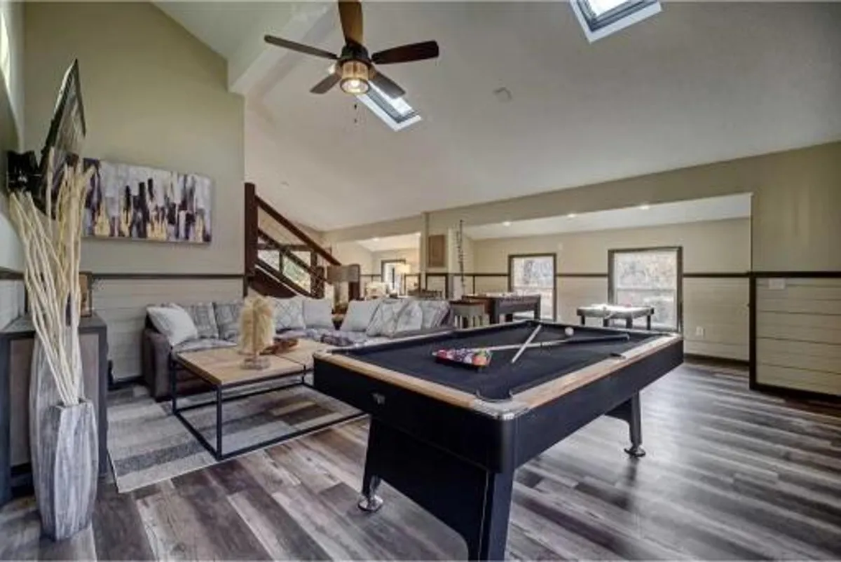 An open-concept entertaining space with a pool table, plush sofa, stylish wall art, and a ceiling fan. Skylights and large windows fill the room with natural light, making it ideal for lively game nights or relaxing with friends and family. The arrangement encourages social interaction and leisurely fun.