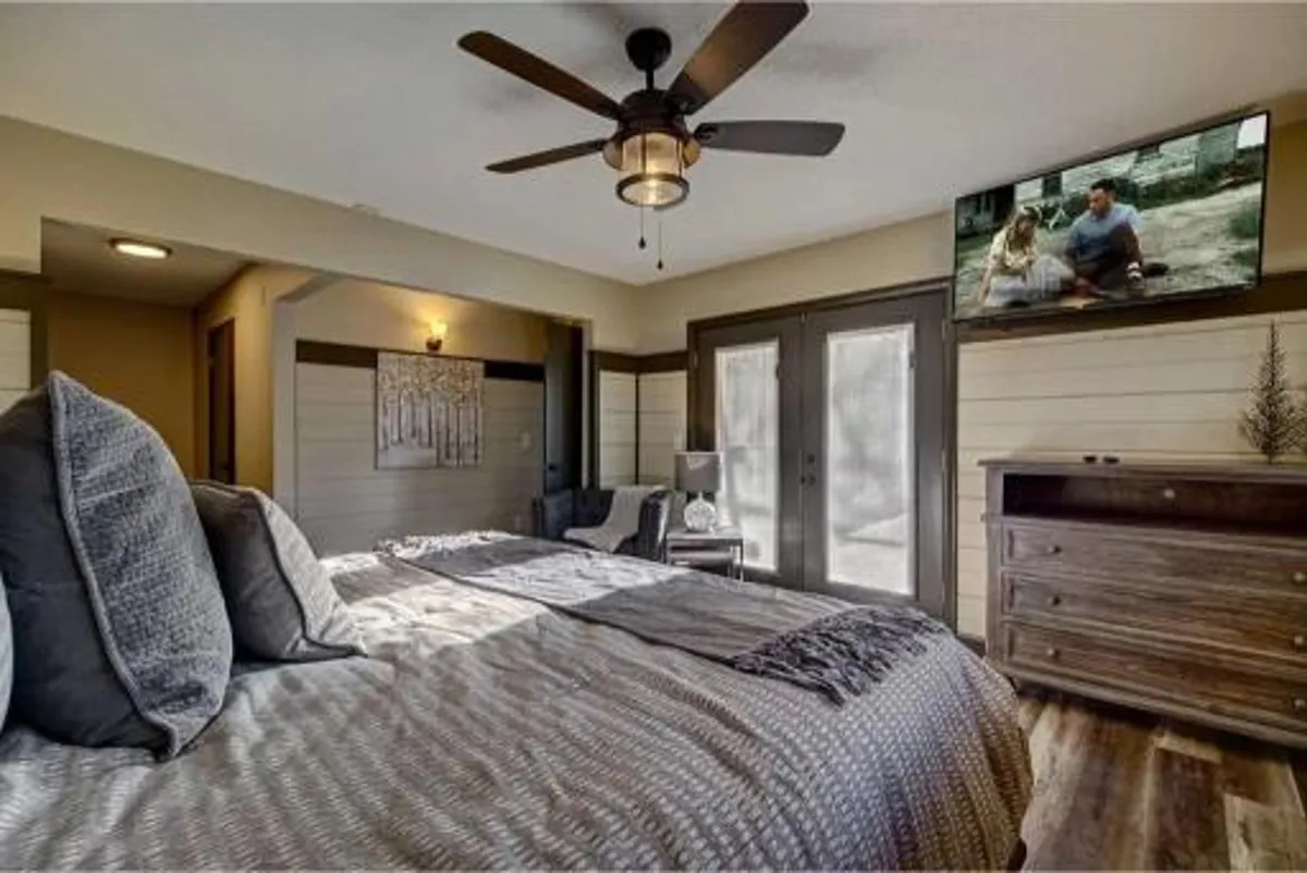 A cozy, inviting bedroom with textured bedding, a ceiling fan, television, and double doors opening to an outdoor area. Sunlight streams in, highlighting the warmth and comfort of the space.